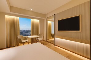 A hotel room with a large bed, a TV mounted on the wall, a dining table with chairs, and a view of the city through the windows. - Lemon Tree Premier, Jabalpur (Jabalpur)