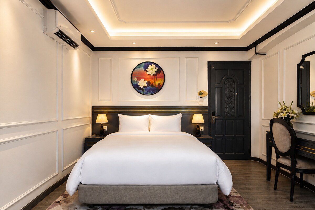 Quarto de hotel com uma cama grande, duas mesas de cabeceira com abajures, uma porta de madeira escura e um quadro redondo na parede.