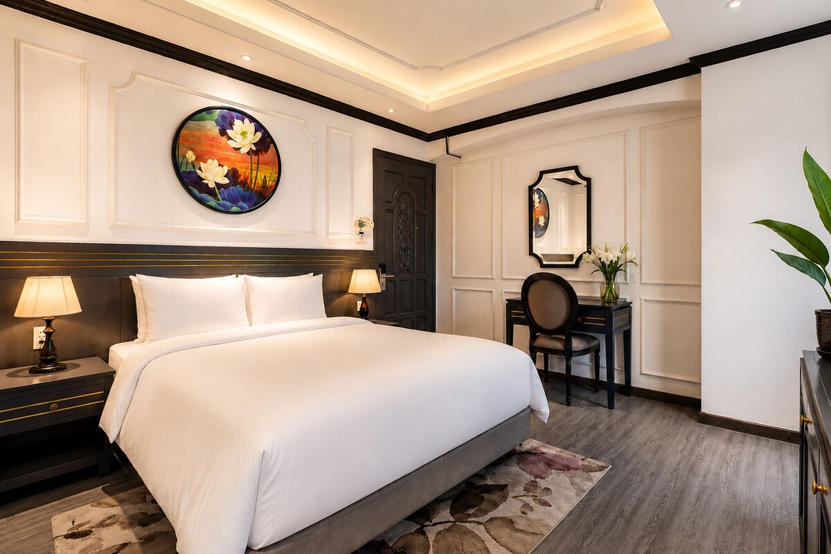 Quarto de hotel com uma cama grande, luminárias de cabeceira, escrivaninha com cadeira, espelho redondo e um quadro na parede.
