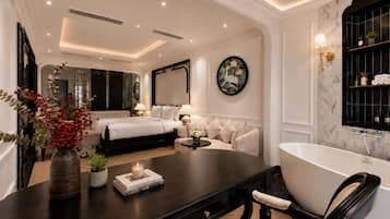 Quarto de hotel moderno com cama grande, área de estar com mesinha de centro, banheira e prateleira fixada na parede com artigos de higiene pessoal.