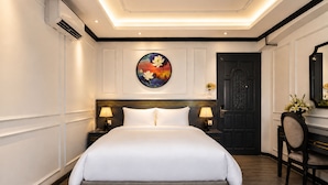 Quarto de hotel com uma cama grande, duas mesas de cabeceira com luminárias, uma porta de madeira escura e um quadro redondo na parede.