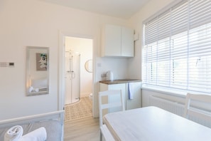 A modern, well-lit room with a shower, a small kitchenette, a dining area, and a large window with blinds. - Bright First Floor Studio with Private Shower Room (Greater London)
