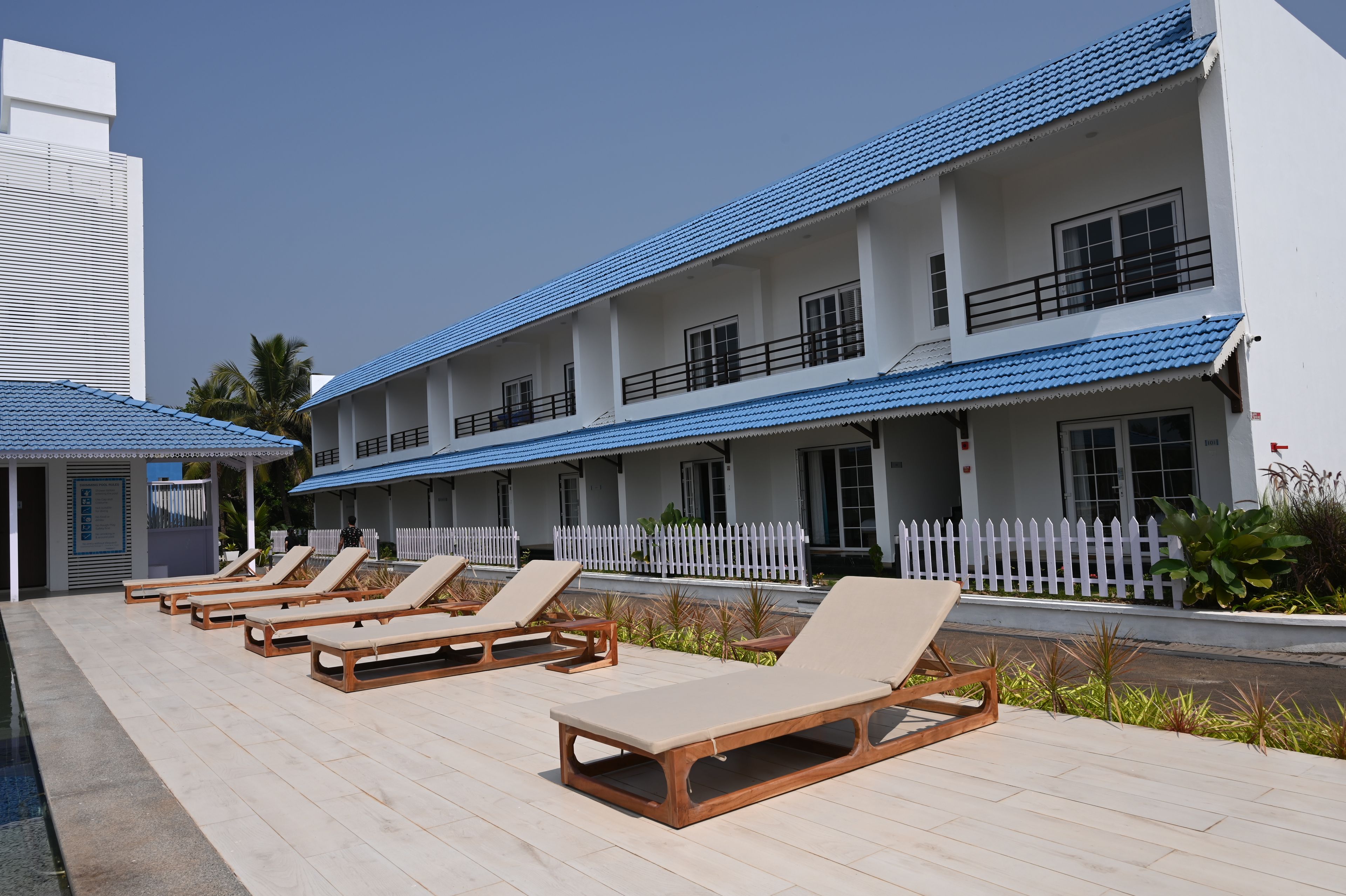 A hotel with multiple floors, a pool area with lounge chairs, and a white picket fence.