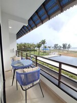 Un balcón con una mesa de vidrio y sillas de mimbre azules, con vista a una playa con palmeras y cielo despejado.