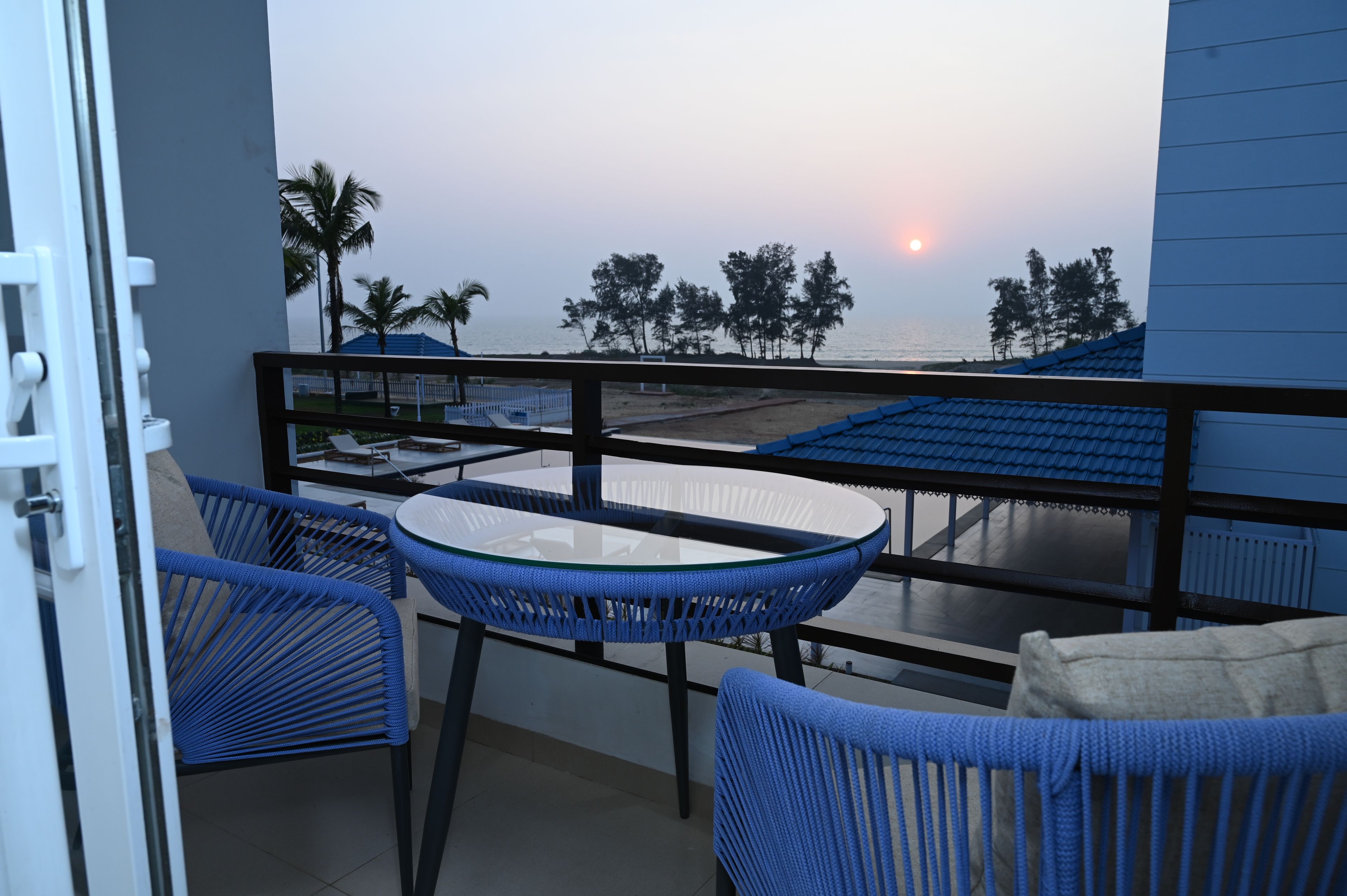 A balcony with a table and chairs, overlooking a beach with palm trees and a sunset.