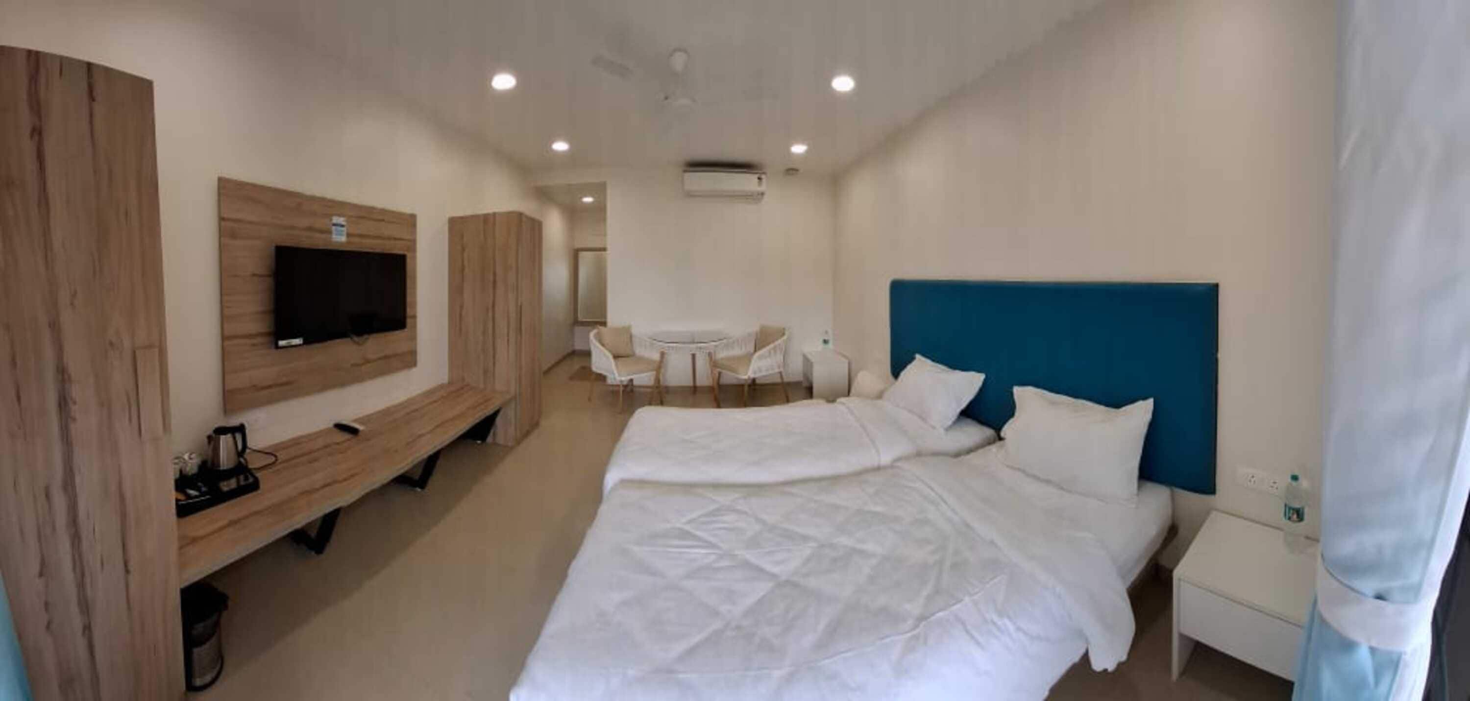 A hotel room with a bed, a TV mounted on the wall, a small table with chairs, and a window with curtains.