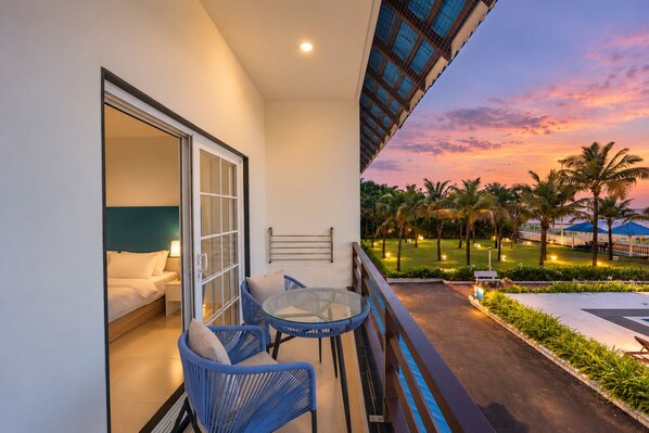 A balcony with a bed, a table, and chairs, overlooking a landscaped area with palm trees and a sunset view.