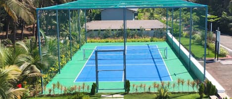 Una cancha de tenis al aire libre con red verde, rodeada por una cerca de metal y exuberante vegetación tropical.