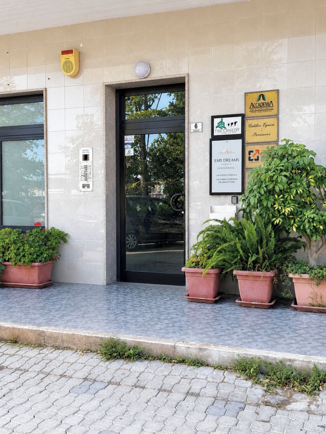 Ein Gebäudeeingang mit einer Kachelfassade, einer Tür, einem Rauchmelder, einem gerahmten Schild und Topfpflanzen.