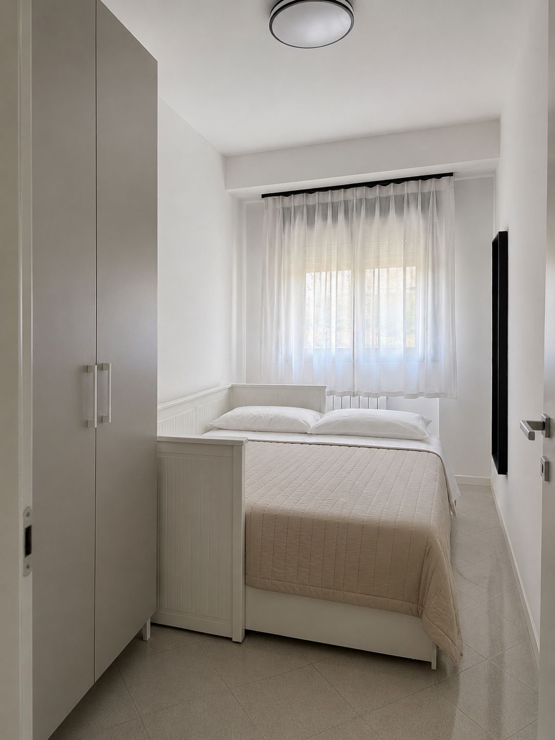 A bedroom with a bed, a window with sheer curtains, a radiator, and a wall-mounted TV.