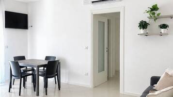 Een moderne woonkamer met een grijze bank, een eettafel met stoelen en een aan de muur bevestigde televisie.
