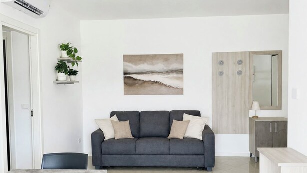 Un espacio de estar moderno con comedor, un sofá gris con cojines, un cuadro en la pared y una cocina con armarios.