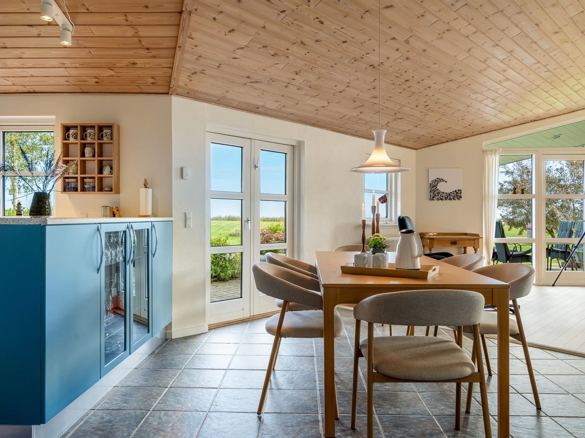 Une cuisine moderne avec un îlot central bleu, un coin repas avec une table et des chaises en bois, et une vue sur l’extérieur grâce à de grandes fenêtres.