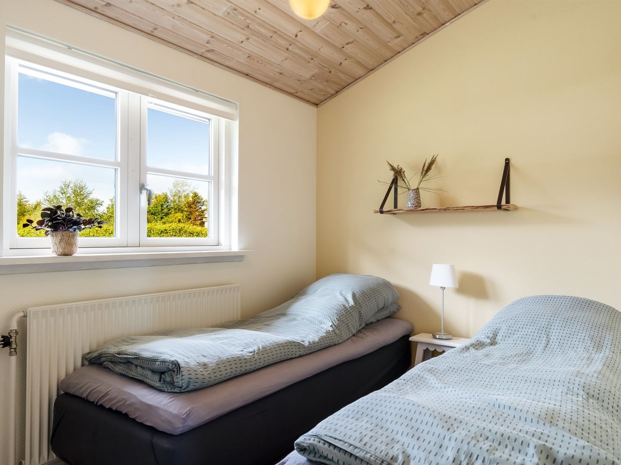 Une chambre avec deux lits, une fenêtre, un radiateur, une table de chevet avec une lampe et une étagère avec des objets décoratifs.