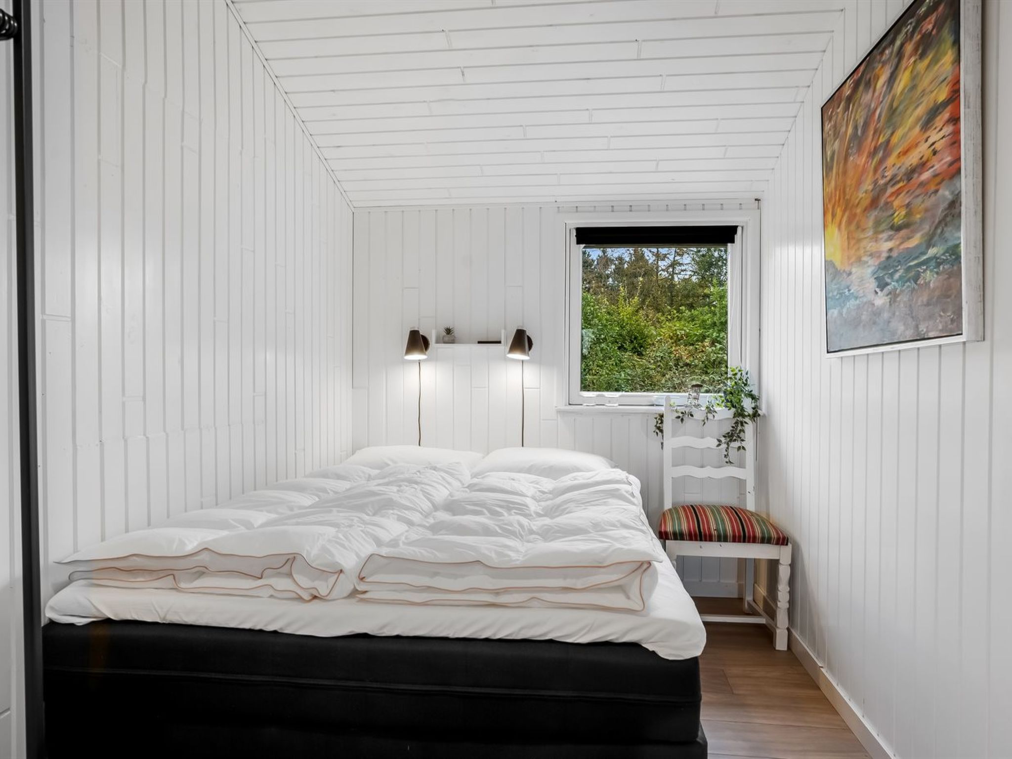 A bedroom with a bed, bedside lamps, a window with a view, and a painting on the wall.