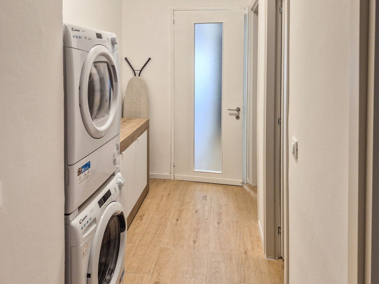 Uma lavanderia com máquinas de lavar empilhadas, uma porta com vidro jateado e piso de madeira.