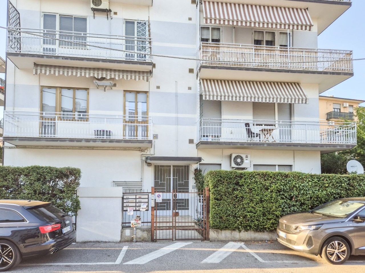 Um edifício residencial de vários andares com varandas, entrada com portão e carros estacionados.