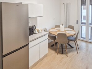 Eine kompakte Küche mit Kühlschrank, Mikrowelle und Kaffeemaschine. Ein runder Esstisch mit Stühlen und ein Fenster mit Jalousien sind vorhanden.