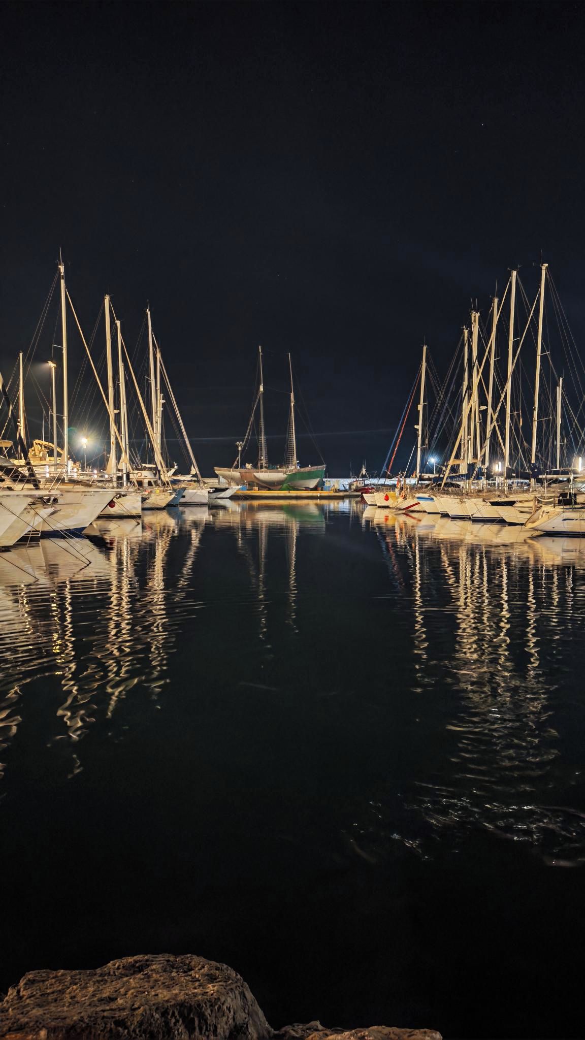 Una marina de noche con varias embarcaciones amarradas, agua tranquila y un primer plano rocoso.