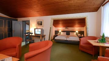 Habitación de hotel con techo de madera, cama con cabecera de madera, dos sillones tapizados en rojo, una mesita con una botella y un televisor sobre un escritorio.