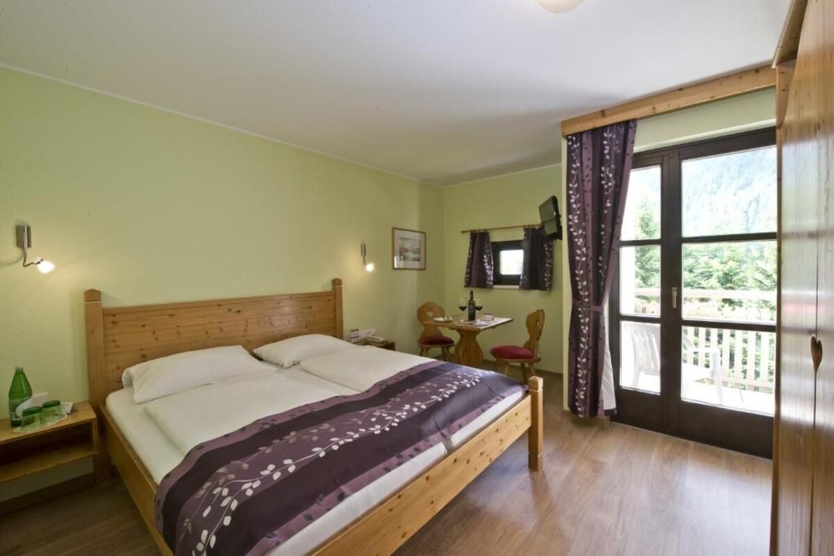 Une chambre avec un lit en bois, une petite table avec des chaises, une fenêtre avec des rideaux et une vue sur la verdure à l’extérieur.