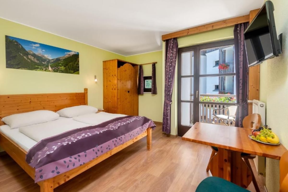 Une chambre avec un lit, un bureau sur lequel se trouve une assiette de fruits, une armoire en bois et une fenêtre avec des rideaux.