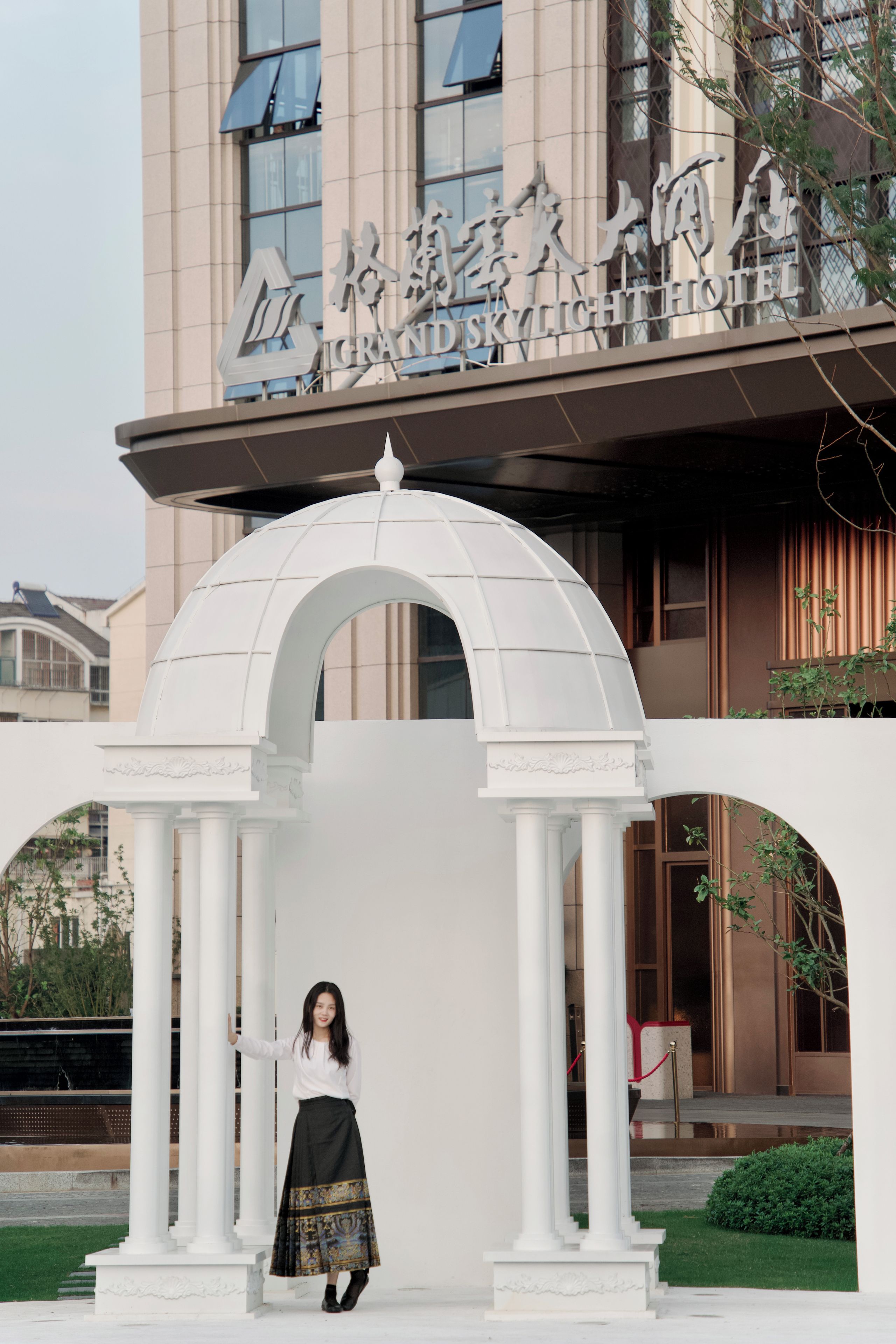 Entrada de hotel con un arco blanco y columnas, un letrero con el nombre del hotel, y una persona parada frente a él.