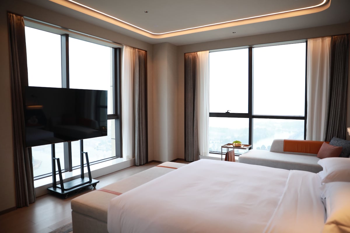 A modern hotel room with a large bed, a smaller bed, a TV, and a view of the city.