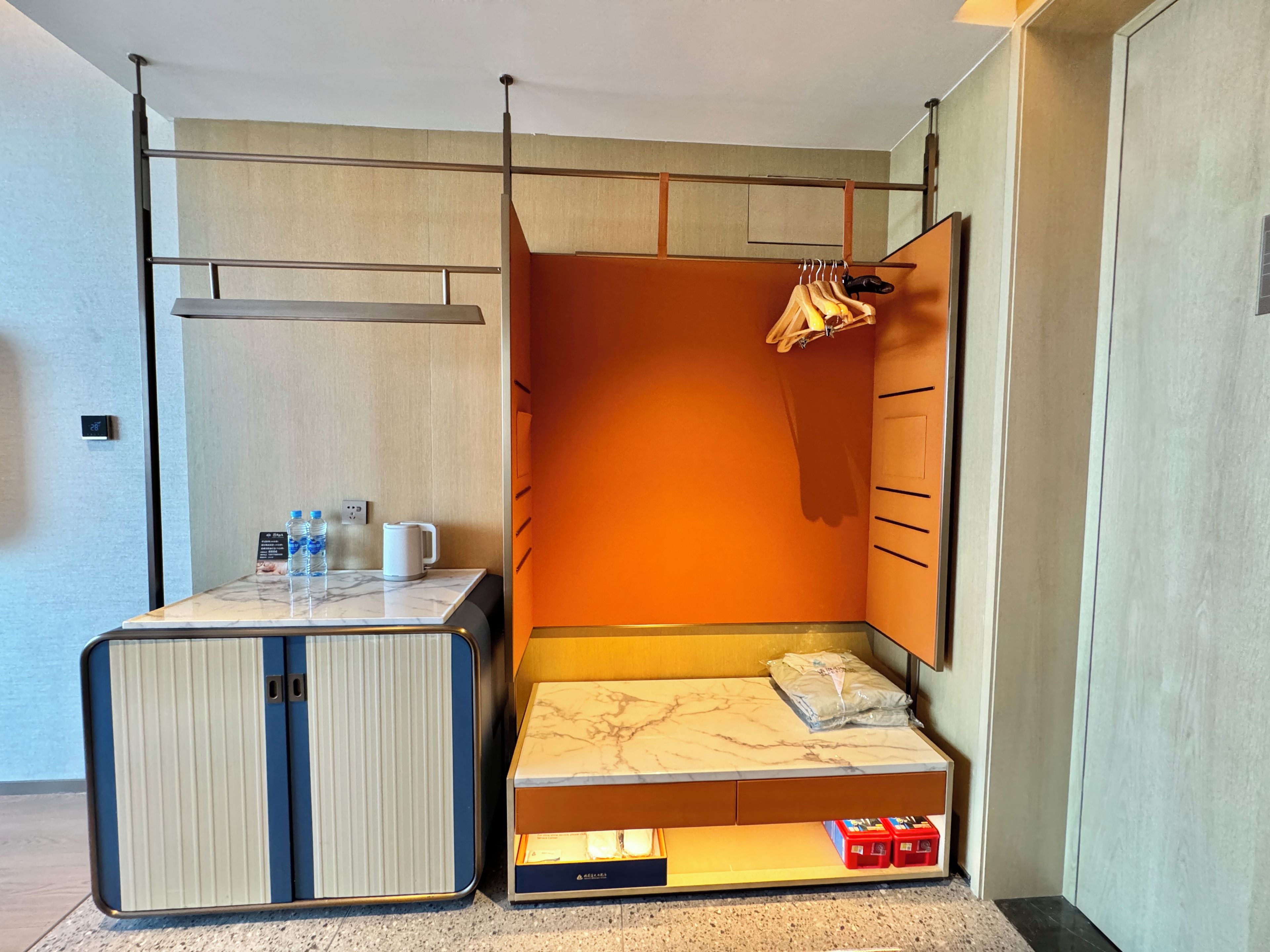 A compact room with a bed, a small table with a kettle and water bottles, and a wardrobe with hanging clothes.