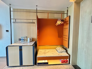 A compact room with a bed, a small table with a kettle and water bottles, and a wardrobe with hanging clothes.