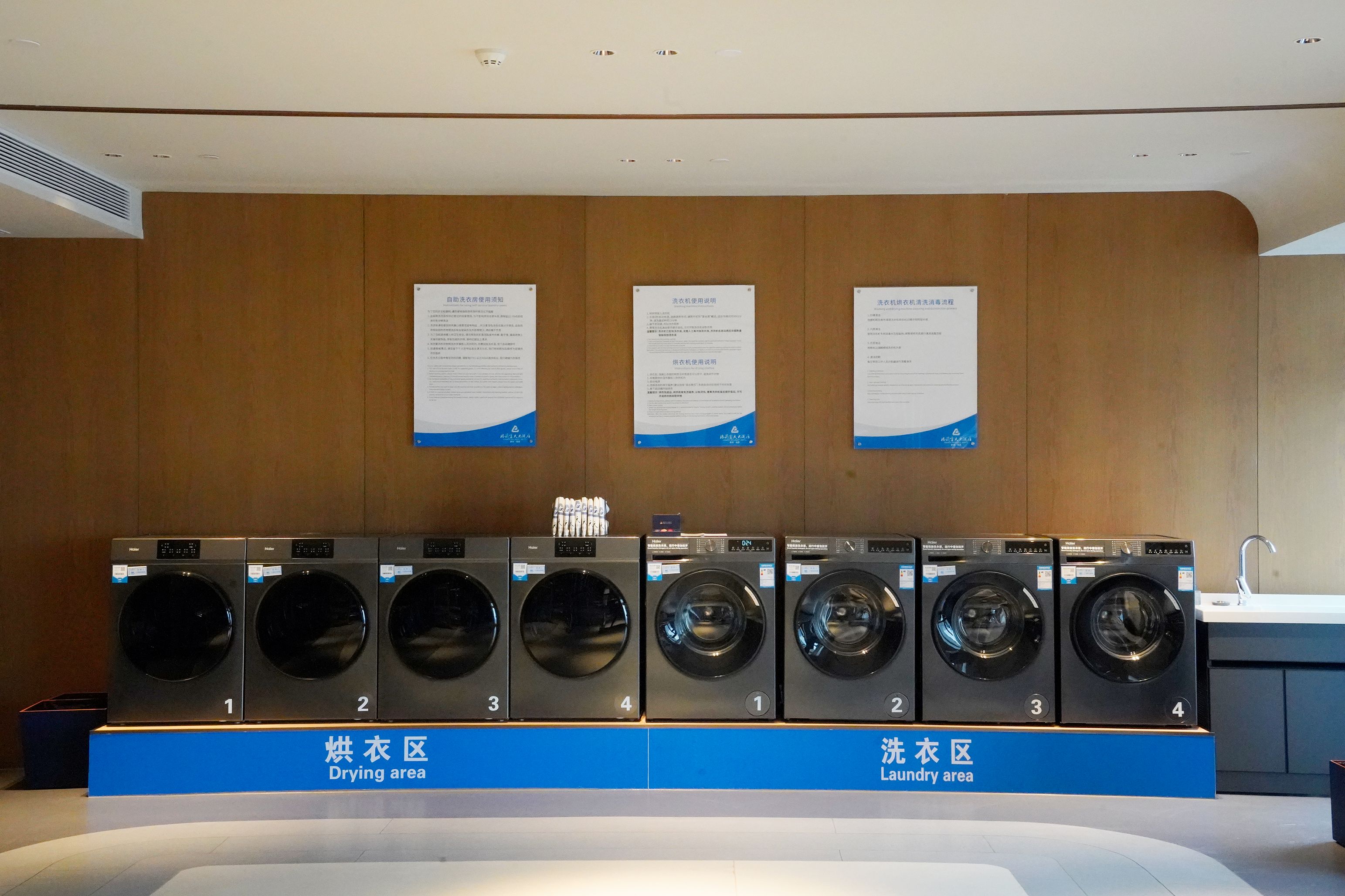 A row of six front-loading washing machines in a commercial setting, with informational posters on the wall above and labels indicating the drying and laundry areas.