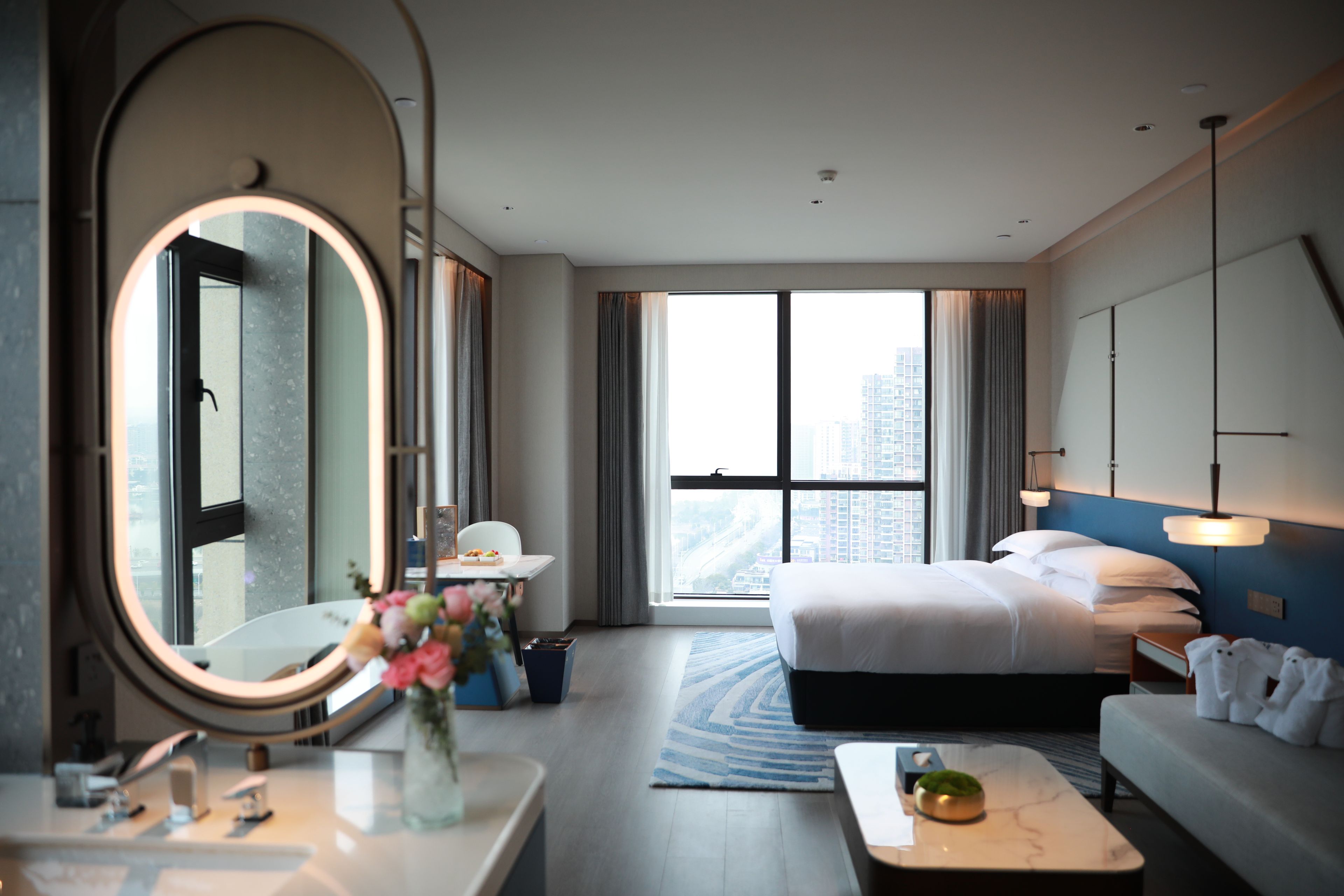 Habitación de hotel con una cama grande, una zona de estar, un espejo redondo, una mesita con un florero y vistas a la ciudad a través de amplias ventanas.
