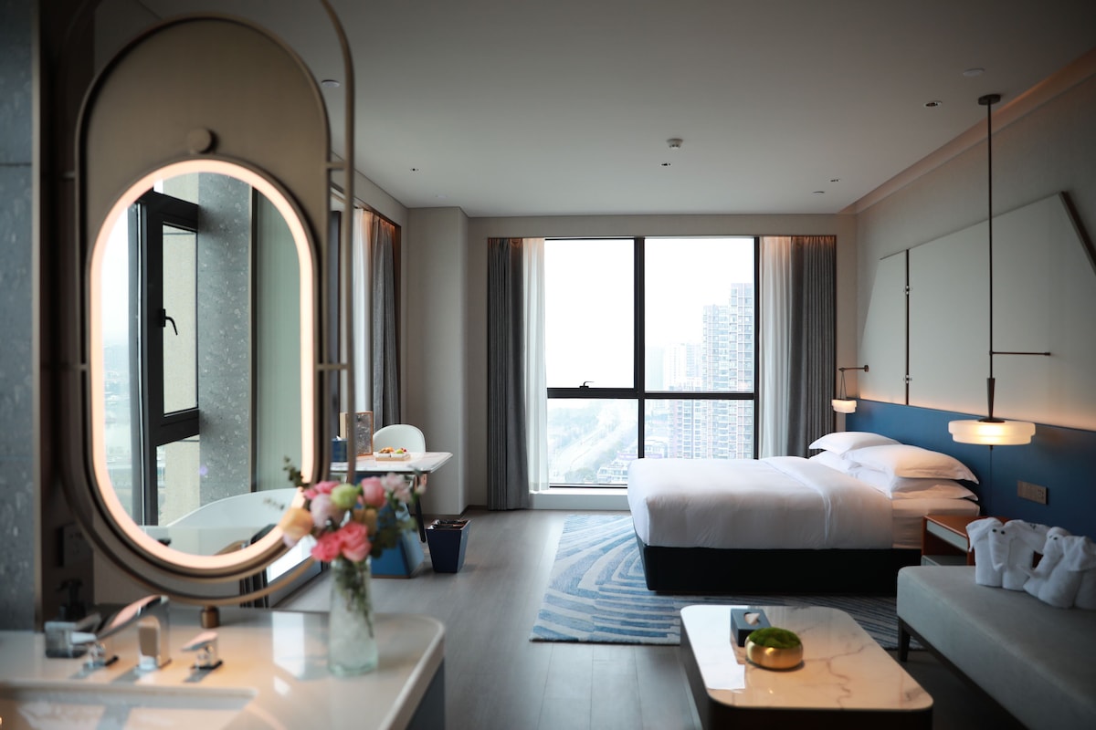 A hotel room with a large bed, a sitting area, a round mirror, a small table with a vase of flowers, and a view of the city through large windows.