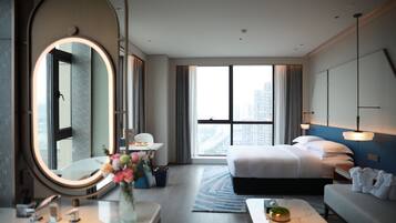 Habitación de hotel con una cama grande, una zona de estar, un espejo redondo, una mesita con un florero y vistas a la ciudad a través de amplias ventanas.