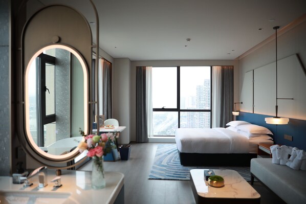 A hotel room with a large bed, a sitting area, a round mirror, a small table with a vase of flowers, and a view of the city through large windows.