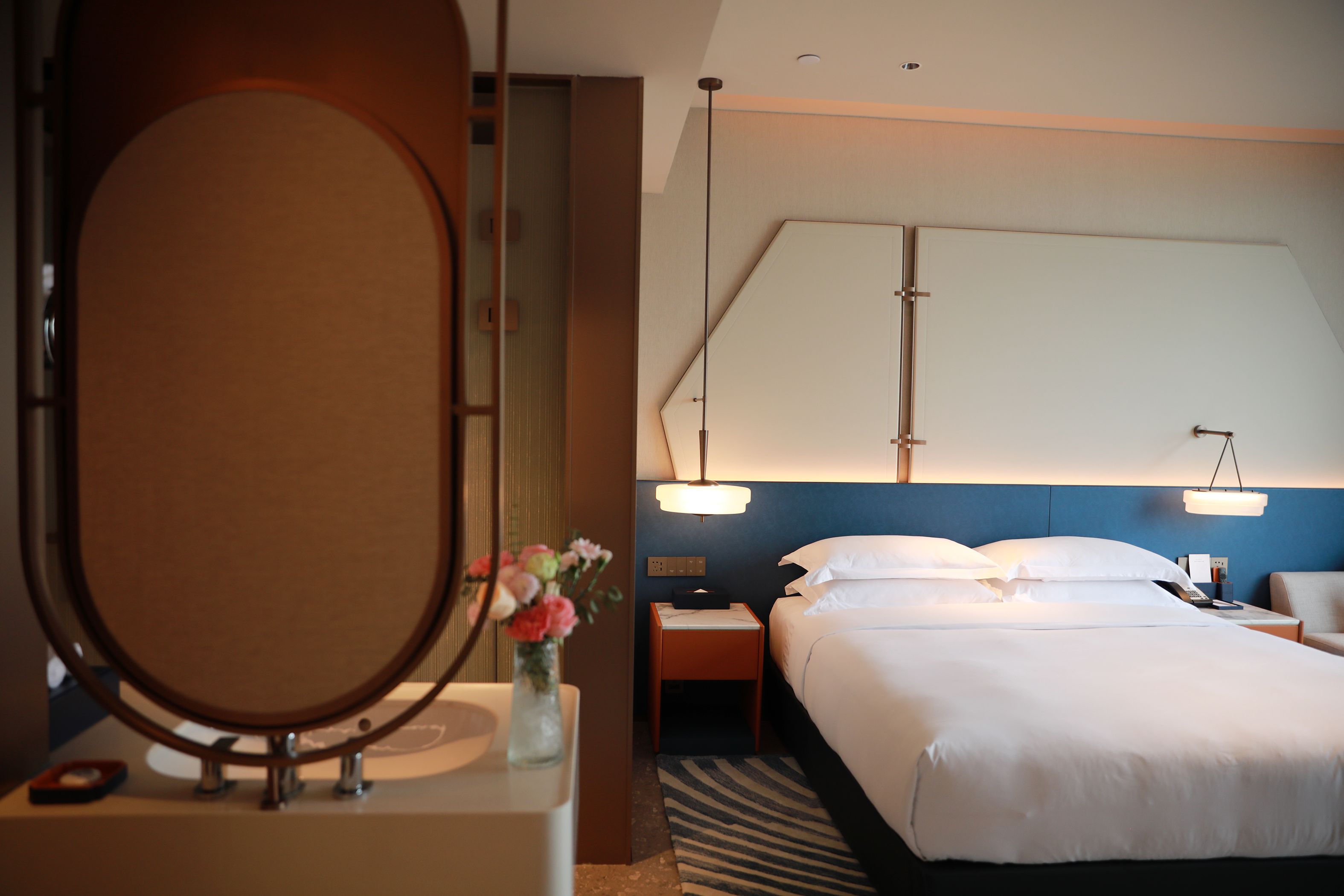 Quarto de hotel com cama grande, luminárias de cabeceira, mesa de apoio, espelho e um vaso com flores.