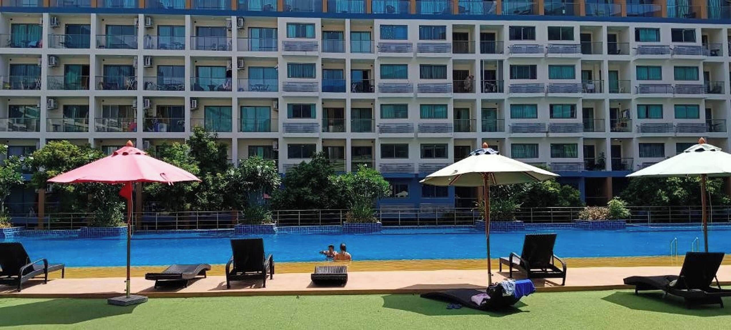 A hotel pool area with lounge chairs, umbrellas, and a building with balconies in the background.
