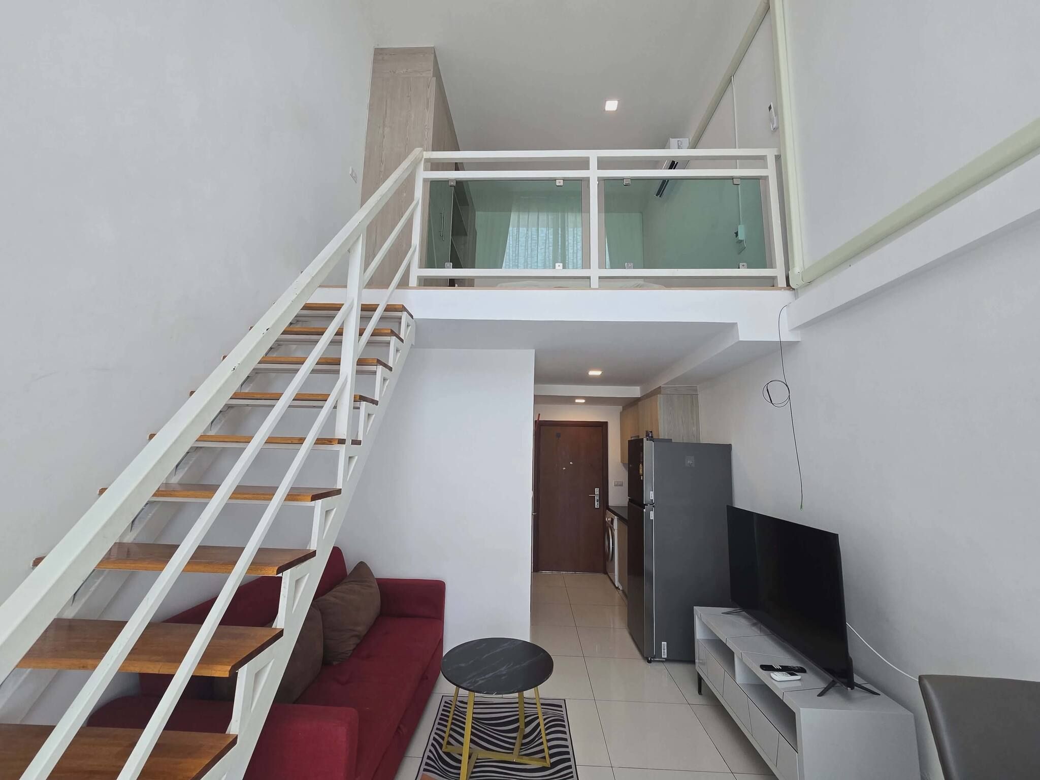 Un appartement moderne avec un escalier menant à une mezzanine, un espace de vie avec un canapé et un téléviseur, et une cuisine équipée d’un réfrigérateur.