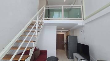 Un appartement moderne avec un escalier menant à une mezzanine, un espace de vie avec un canapé et un téléviseur, et une cuisine équipée d’un réfrigérateur.