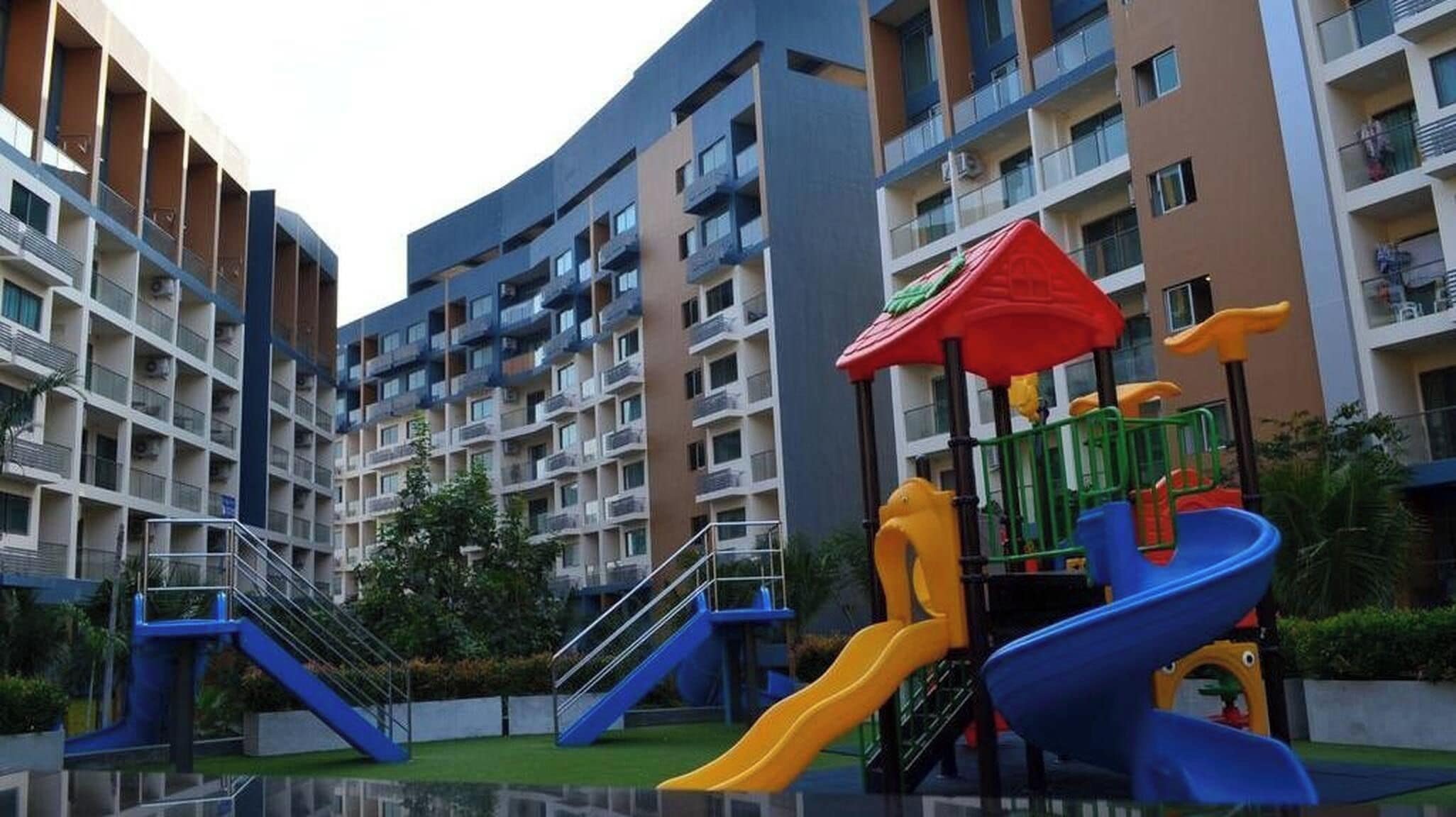 A multi-story residential building with balconies, a playground with slides, and a small water feature.