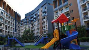 En flervåningsbyggnad med bostäder, balkonger, en lekplats med rutschkanor och en liten vattenfontän.