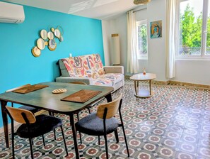 Una habitación con pared azul, piso estampado, una mesa de comedor con sillas, un sofá con una manta colorida y una pequeña mesa redonda con una vela.
