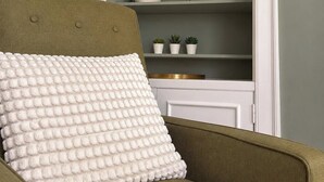 Een kamer met een groene fauteuil, een wit kussen met reliëf en een wit, gebogen wandrek met decoratieve objecten.