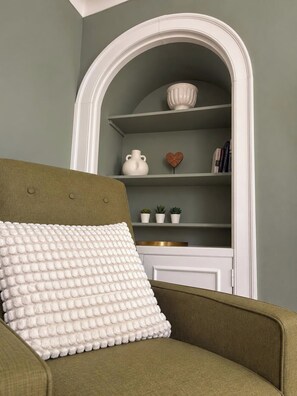 Una habitación con un sillón verde, un cojín blanco texturizado y un estante blanco de arco con objetos decorativos.