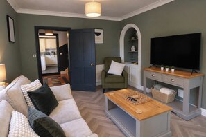 Una sala de estar con un sofá, un televisor sobre un mueble de madera, una puerta que da a una cocina y una silla con cojín.