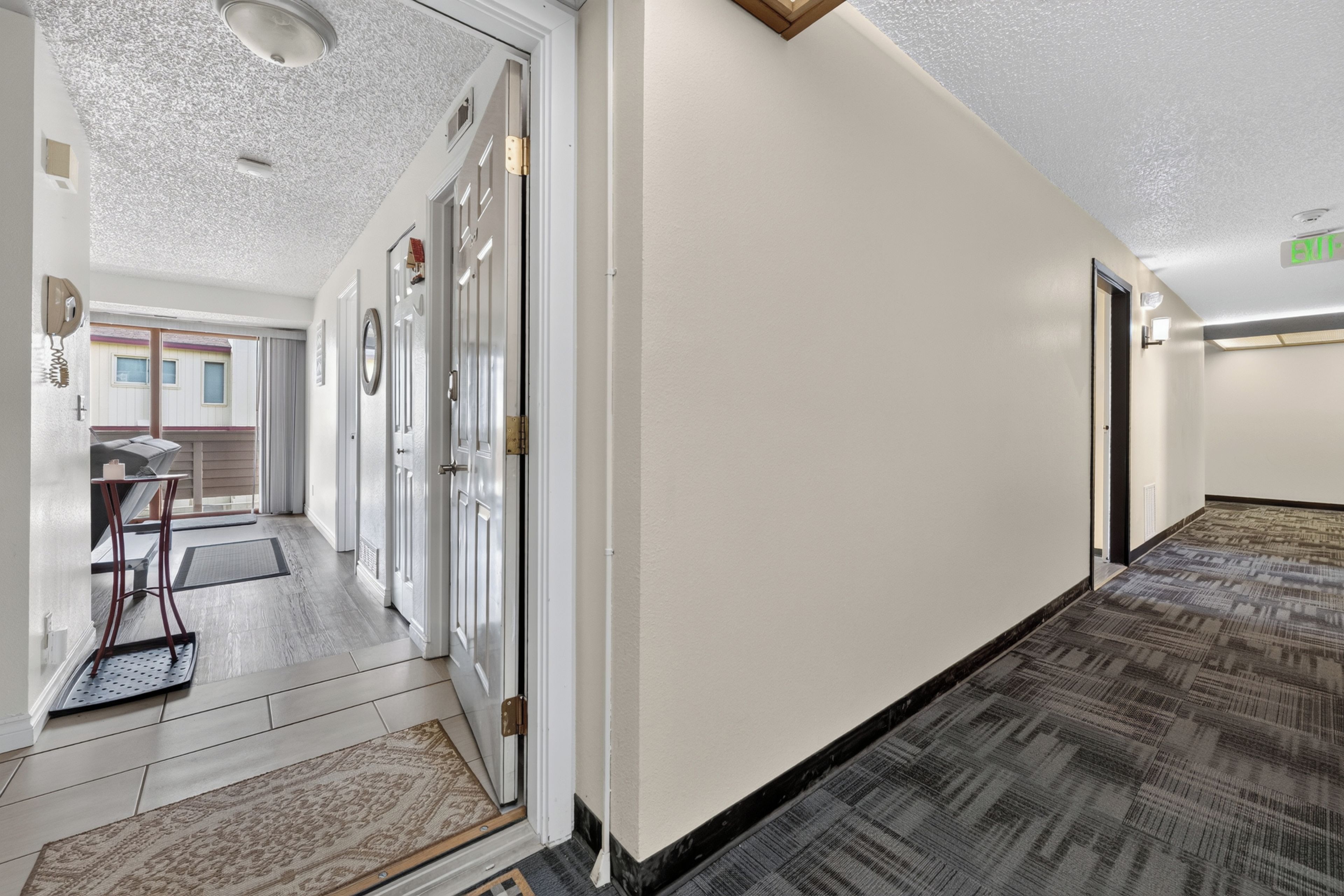 Un couloir d’hôtel avec un tapis à motifs, une porte dotée d’une poignée en laiton et un panneau de signalisation indiquant la sortie, bien visible.