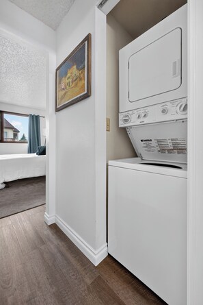 A laundry room with a front-loading washer and dryer, a painting on the wall, and a window with a view of a house. - Remodeled  1-bedroom condo with garage in Anchorage perfect for a relaxing stay (Anchorage)
