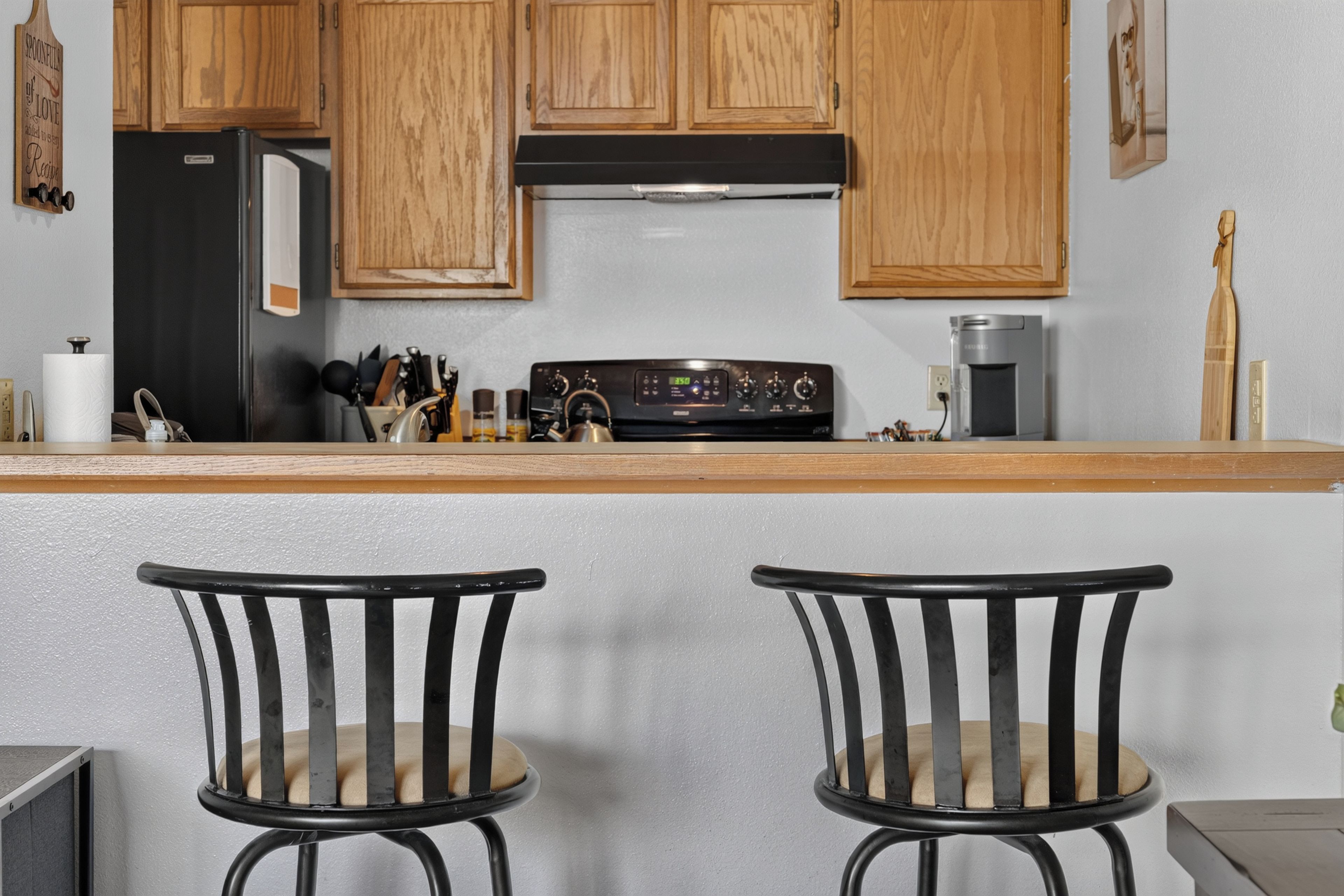 Une cuisine avec des meubles de rangement en bois, un réfrigérateur noir, une cuisinière et deux chaises noires dotées d’assises rembourrées.