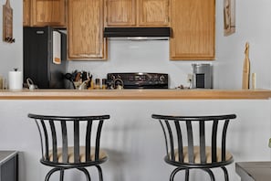 A kitchen with wooden cabinets, a black refrigerator, a stove, and two black chairs with cushioned seats.