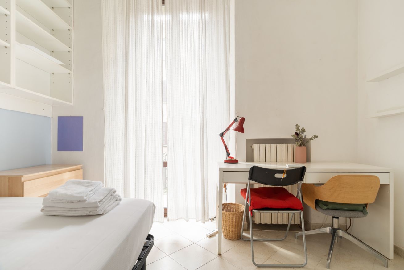 A hotel room with a bed, a desk with a chair, a lamp, and a window with curtains.